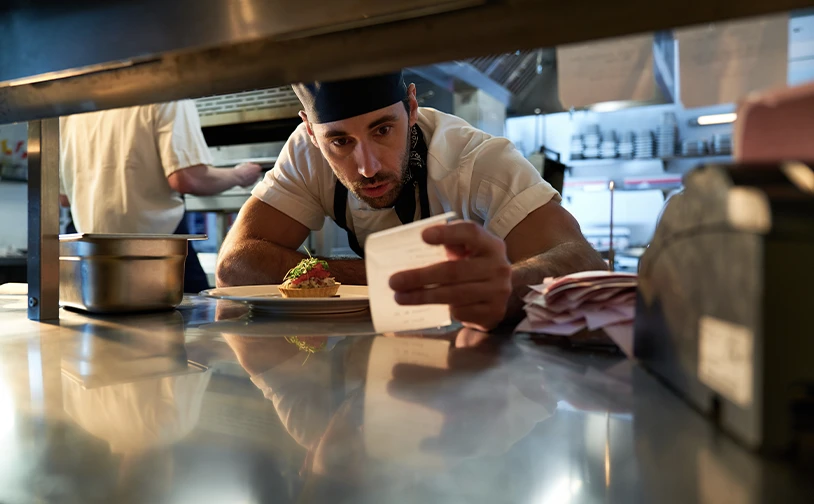 Chef reviewing a printed order ticket at a restaurant pass in a busy commercial kitchen