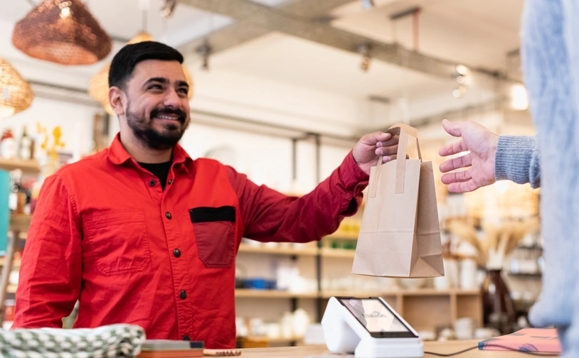 A retail employee hands a paper shopping bag to a customer across the counter